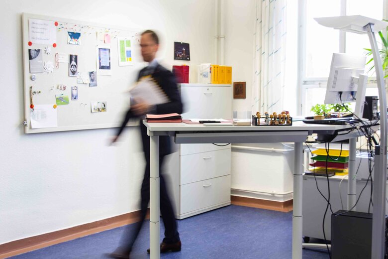 Ein Büro im Altbau der Pädagogischen Hochschule Heidelberg  mit Steh-Sitz-Arbeitstisch im Vordergrund. Ein Mann läuft hinter dem Schreibtisch hervor; er ist lediglich schemenhaft sichtbar.