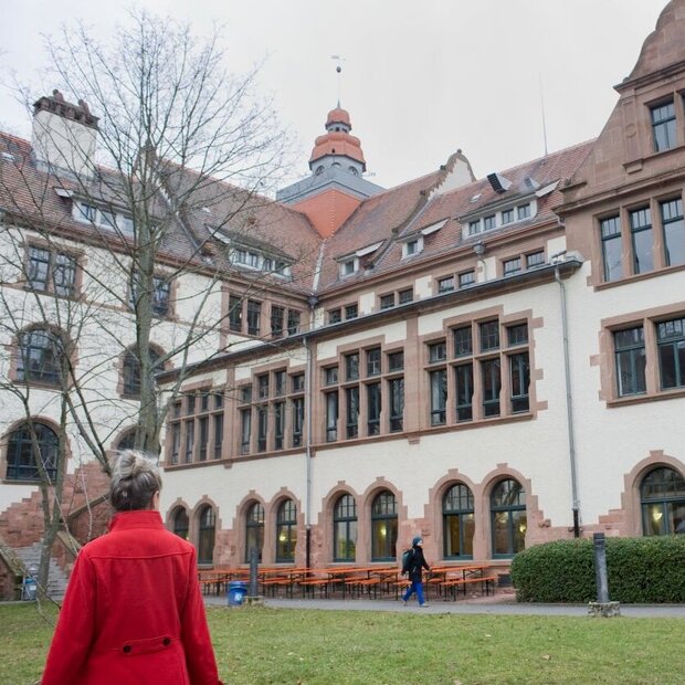 Eine Frau im roten Mantel blickt auf den Altbau. Sie ist lediglich von hinten zu sehen. Das Bild wurde im Innenhof im Winter aufgenommen.
