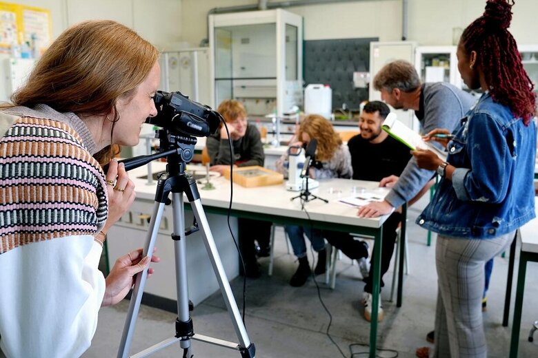 Der Fokus liegt auf einer Forscherin. Sie betrachtet eine Gruppe von Studierenden und einen Professor durch die Kamera. Das Bild wurde in einem Labor der Pädagogischen Hochschule Heidelberg aufgenommen.