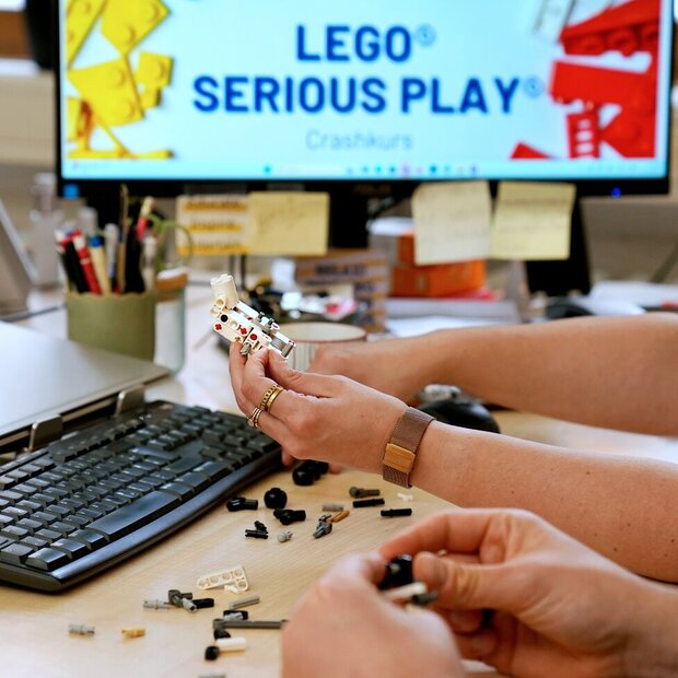 Der Fokus liegt auf den Händern von zwei Personen, die mit Lego etwas bauen. Die Personen sitzen im Büro am Schreibtisch. Auf dem PC-Bildschirm ist "Lego Serious Play" zu lesen.